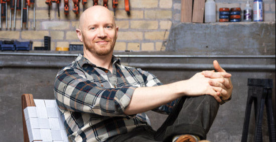A man sits in a modern wooden armchair with a white woven seat and backrest, posed in a workshop with tools hanging on a brick wall behind him.