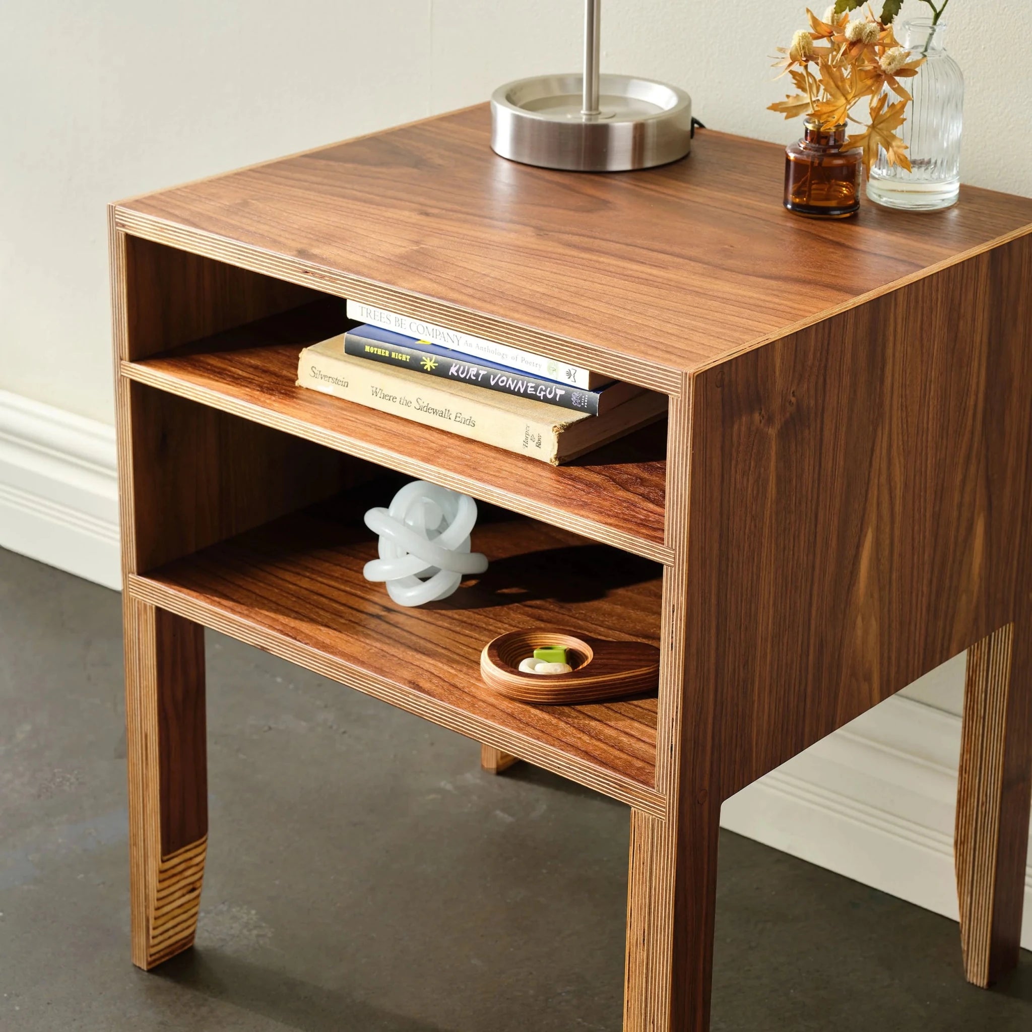 Angled view of walnut open-shelf nightstand with stacked books, lamp and glass knot