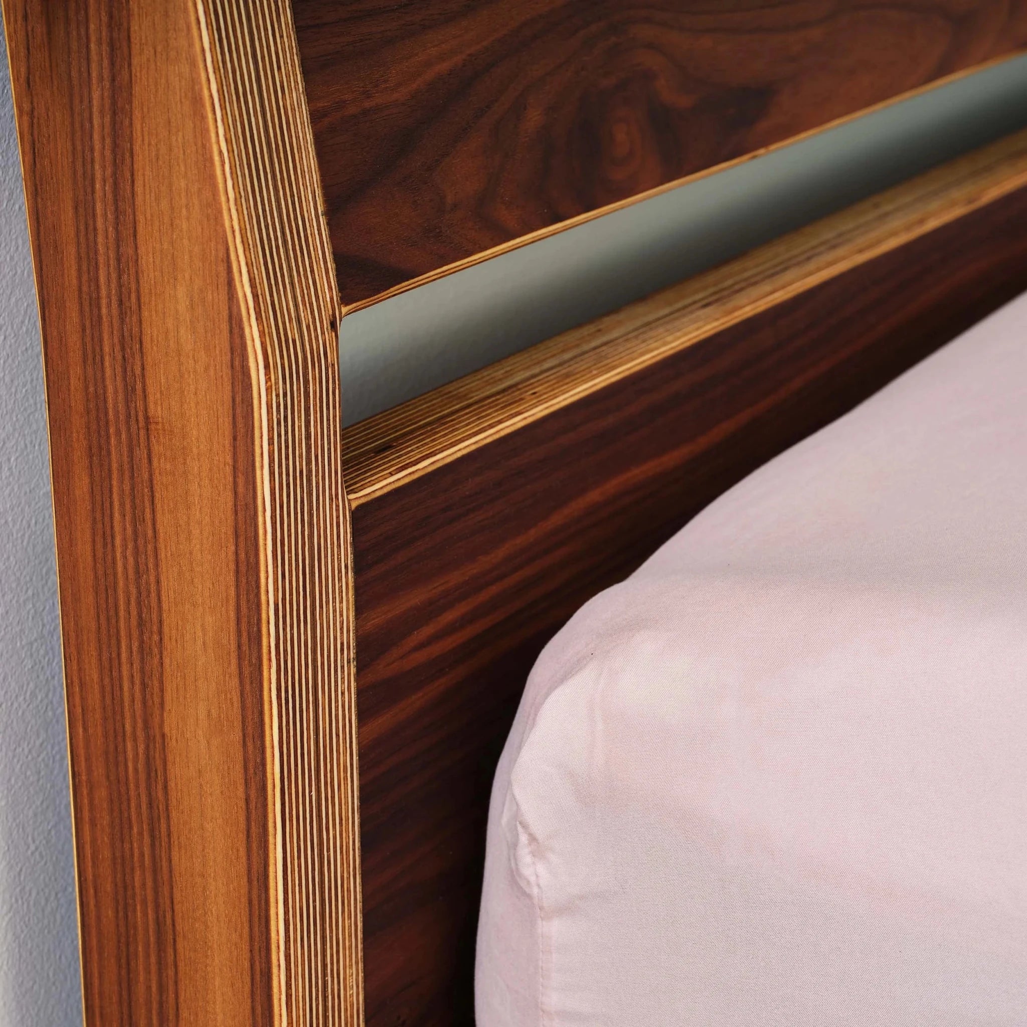 Close-up walnut bed headboard joinery, layered birch edges, pink pillow beside