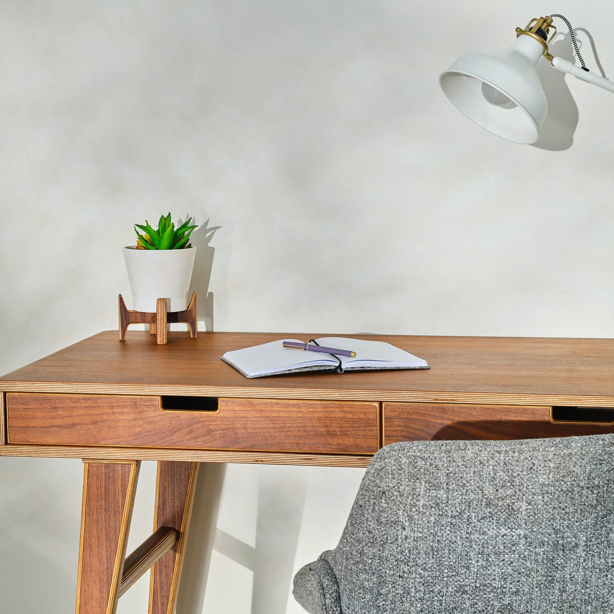 Walnut desk with plywood-edge drawers, plant stand holding succulent, notebook, gray chair, white lamp