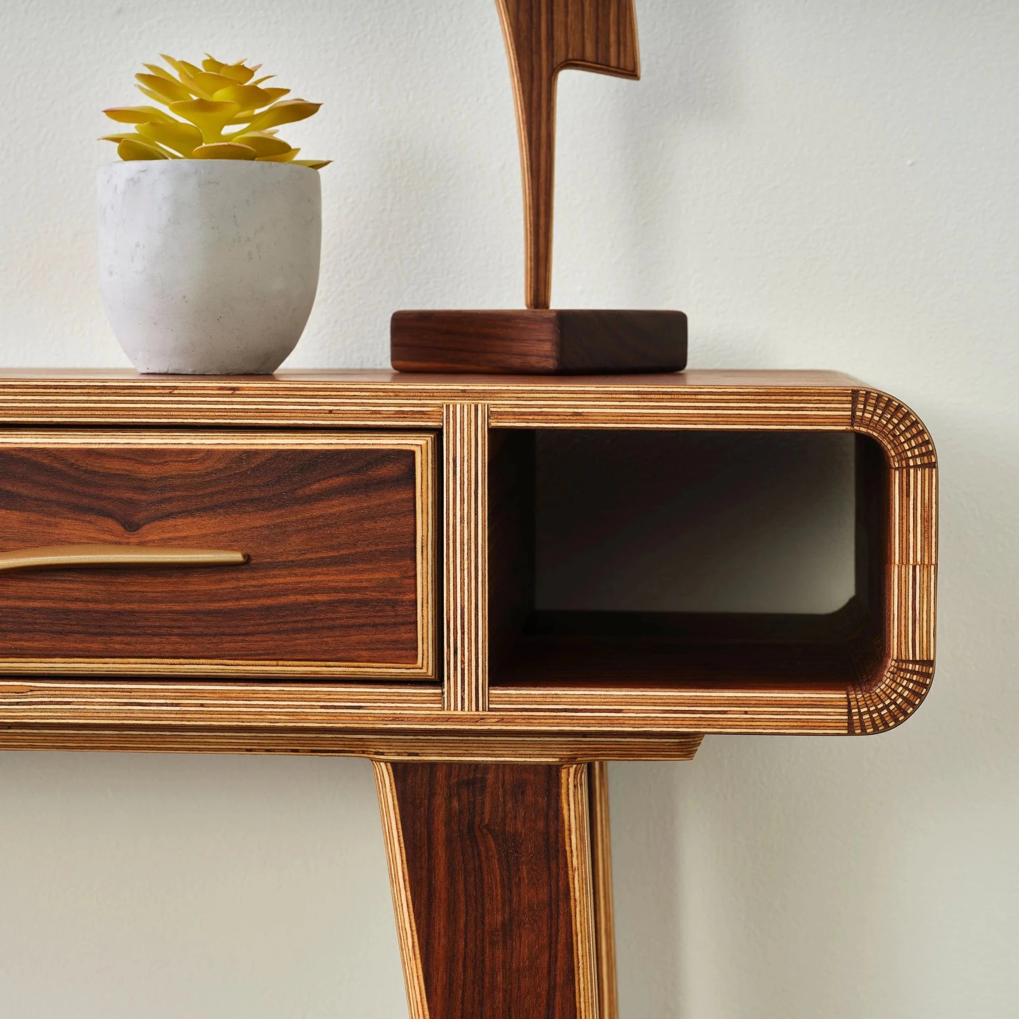 Partial angled view of walnut entry table, brass drawer pull, open cubby, succulent, sculpture
