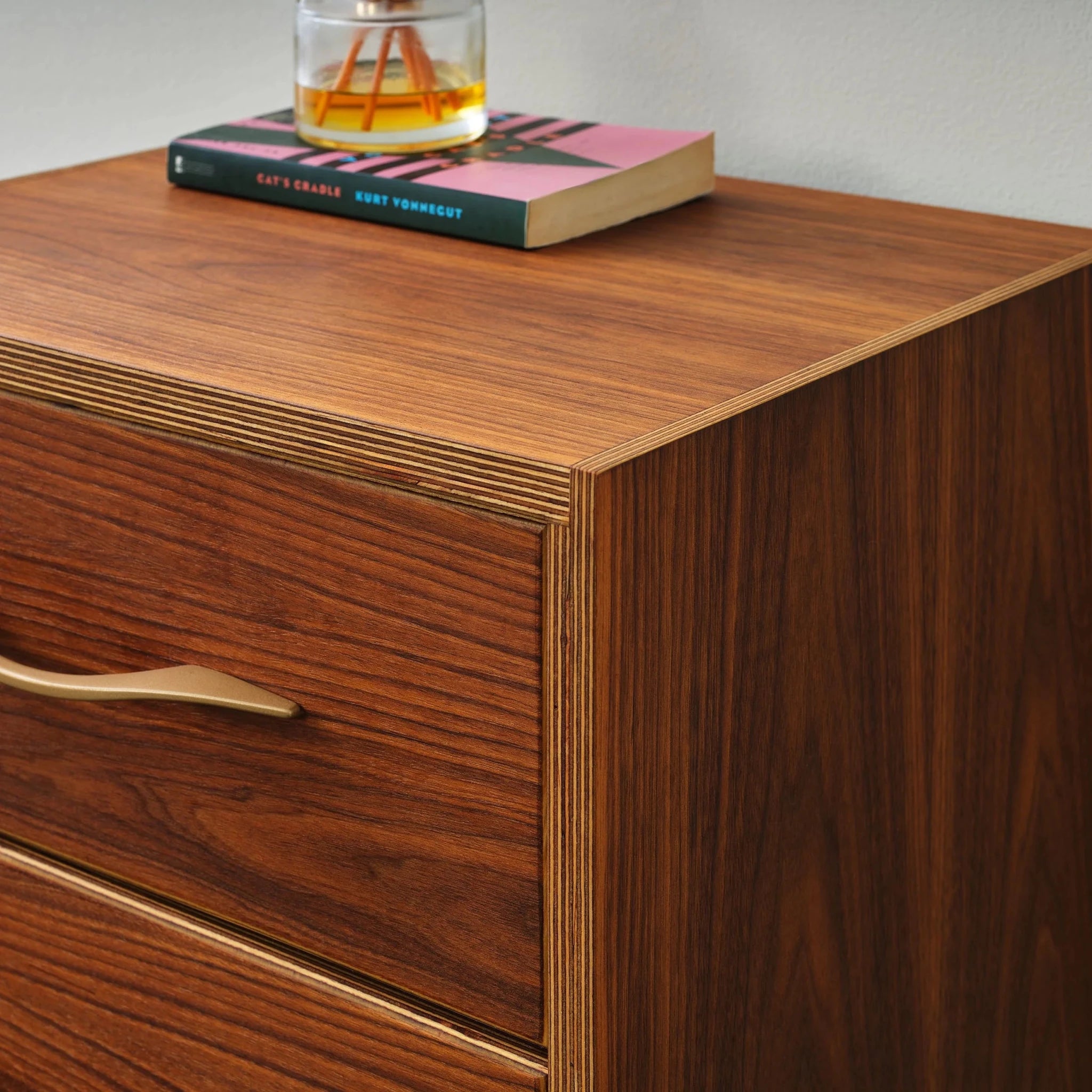 Close-up of walnut nightstand corner, layered plywood edge, book and whiskey glass on top