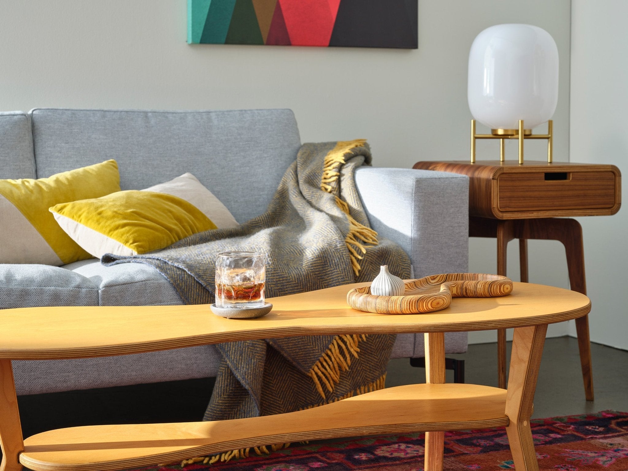Close-up of light birch cocktail table with whiskey glass, gray sofa and yellow pillows behind