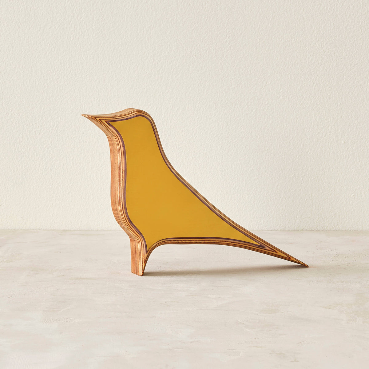A modern bird-shaped decorative sculpture made of layered wood with a matte yellow laminate in a neutral space.