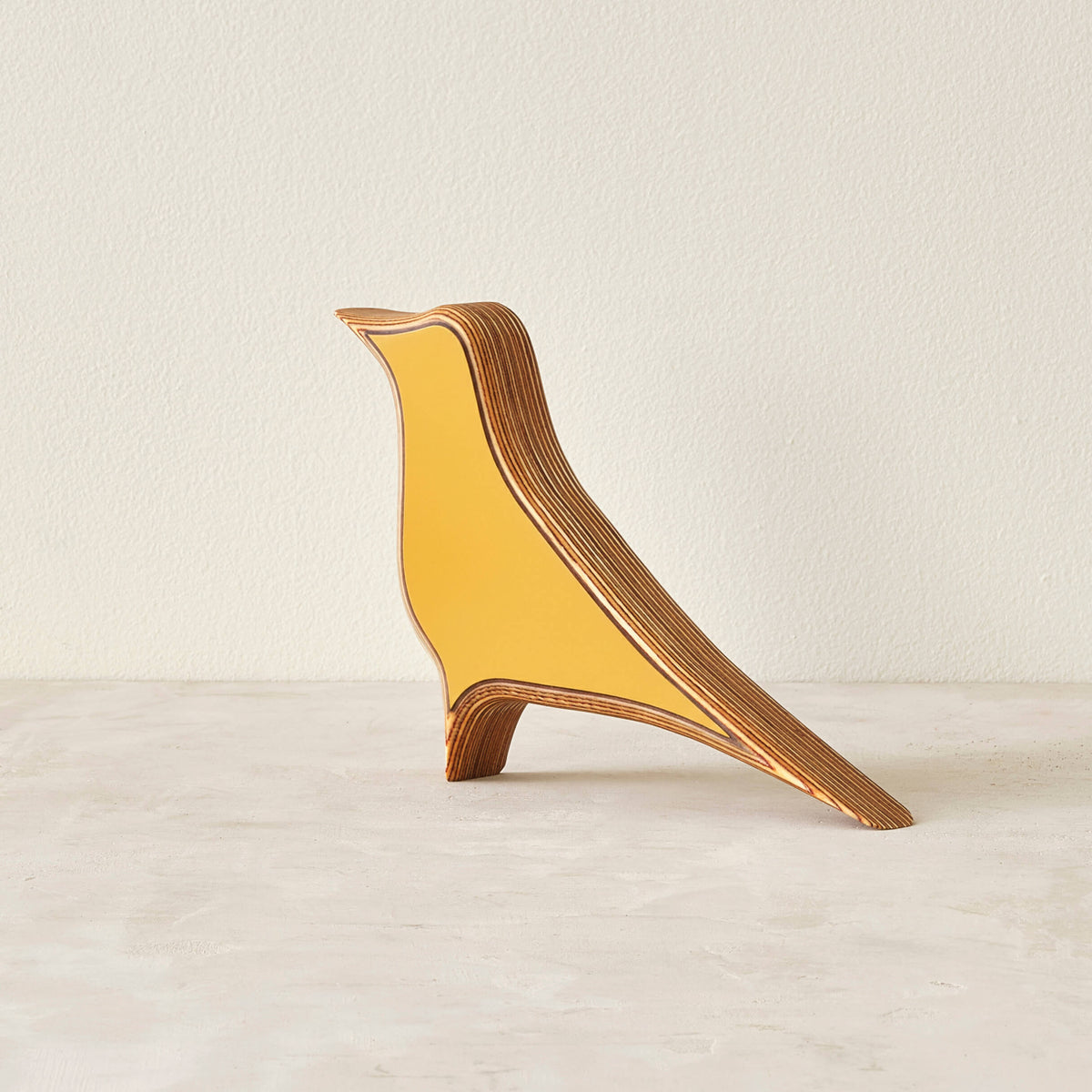 A modern bird-shaped decorative sculpture made of layered wood with a matte yellow laminate in a neutral space.