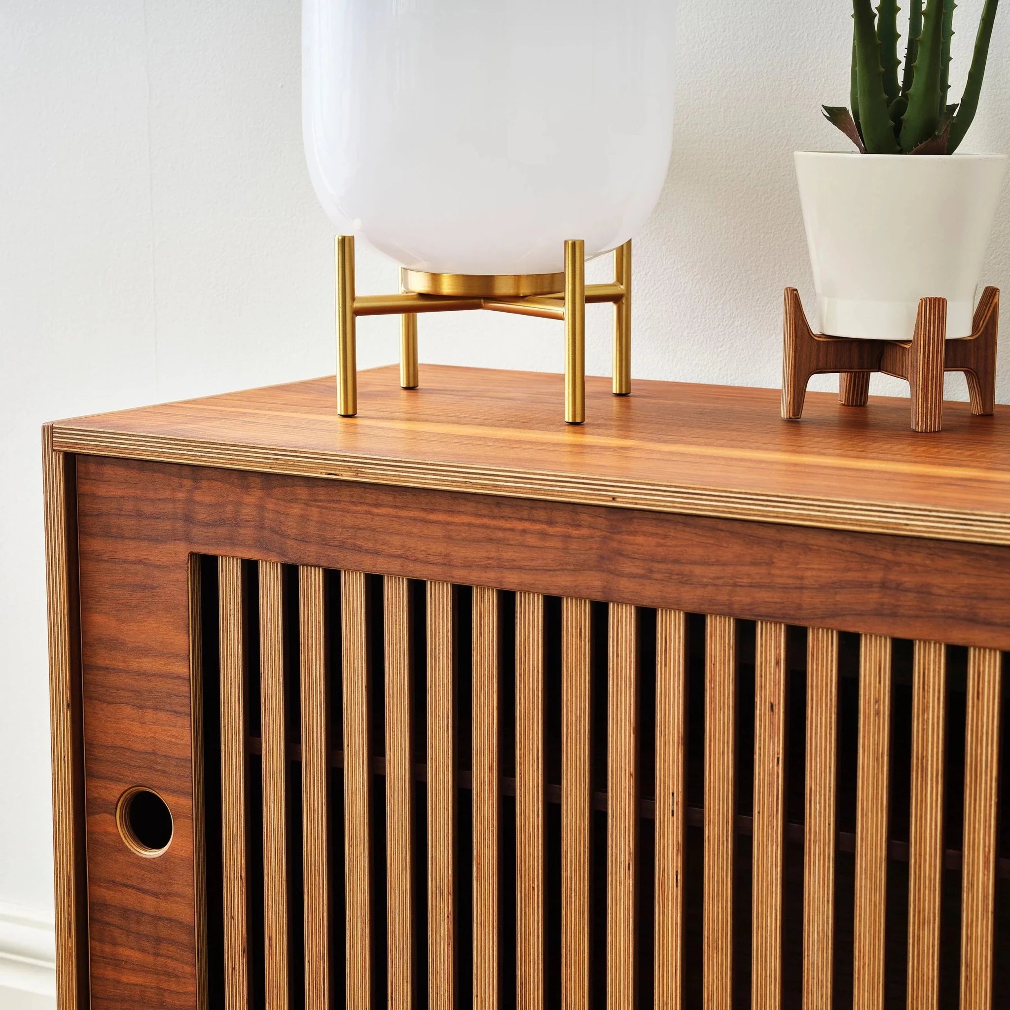 close up of a walnut media console with slatted doors and a plant and lamp on top