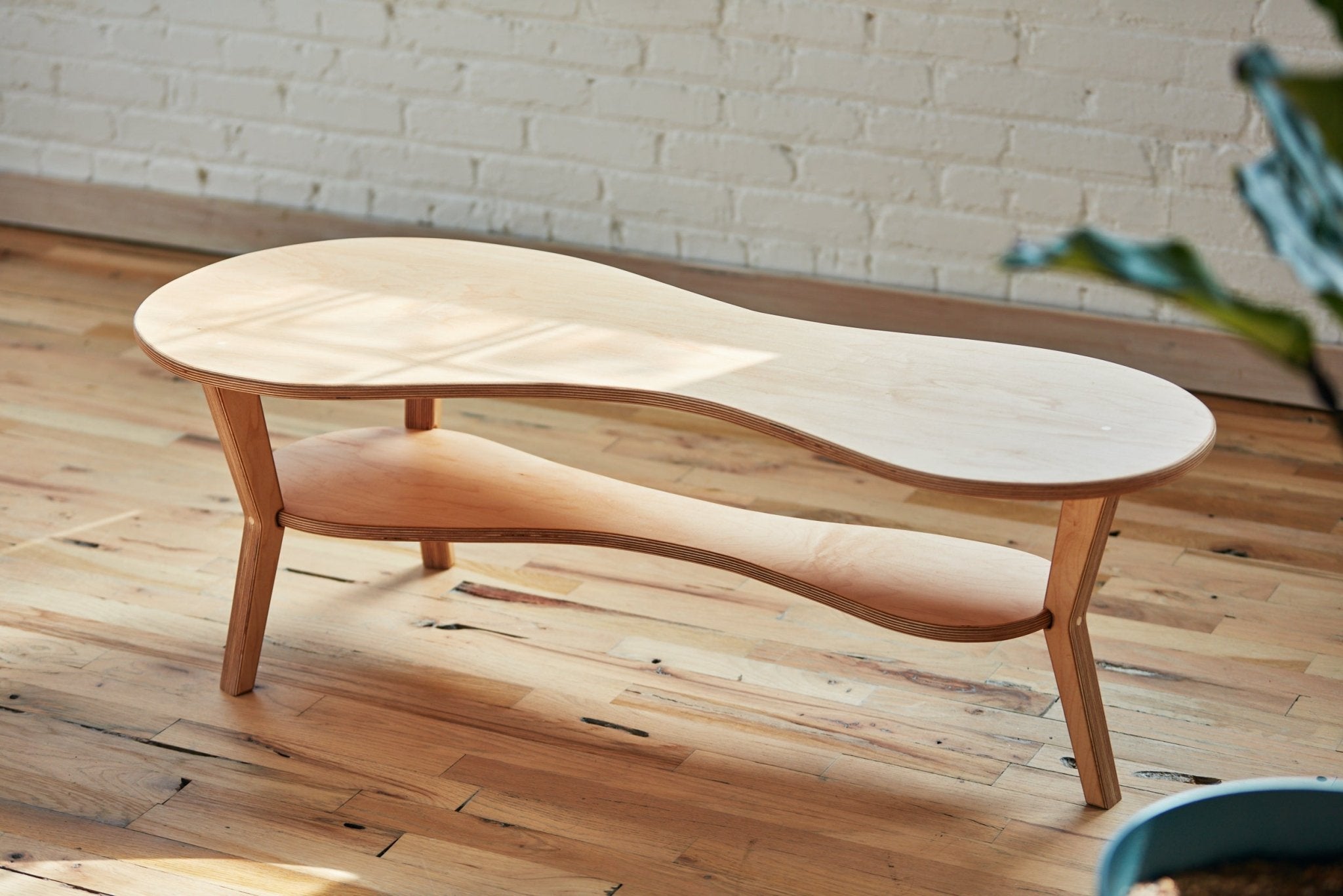Light maple amoeba cocktail table with lower shelf, hardwood floor, white brick wall, studio shot