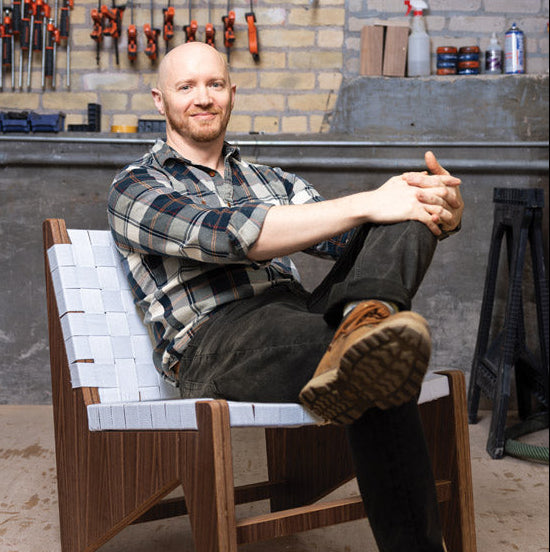 A man sits in a modern wooden armchair with a white woven seat and backrest, posed in a workshop with tools hanging on a brick wall behind him.