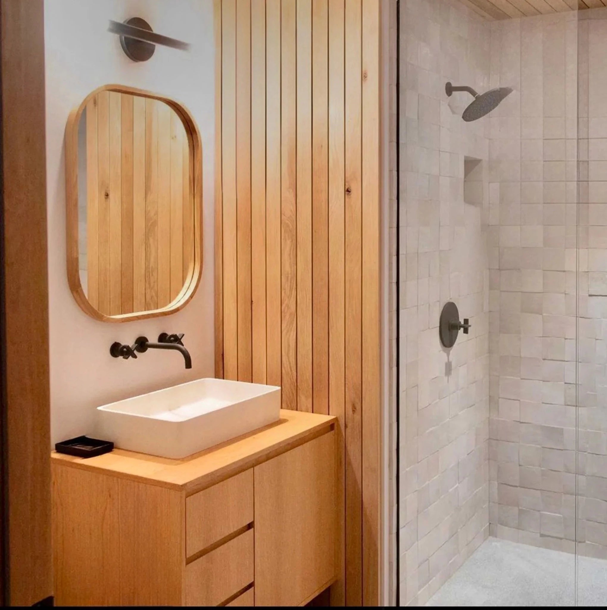 Light oak vanity, white vessel sink, black wall faucet, oval mirror, tiled shower