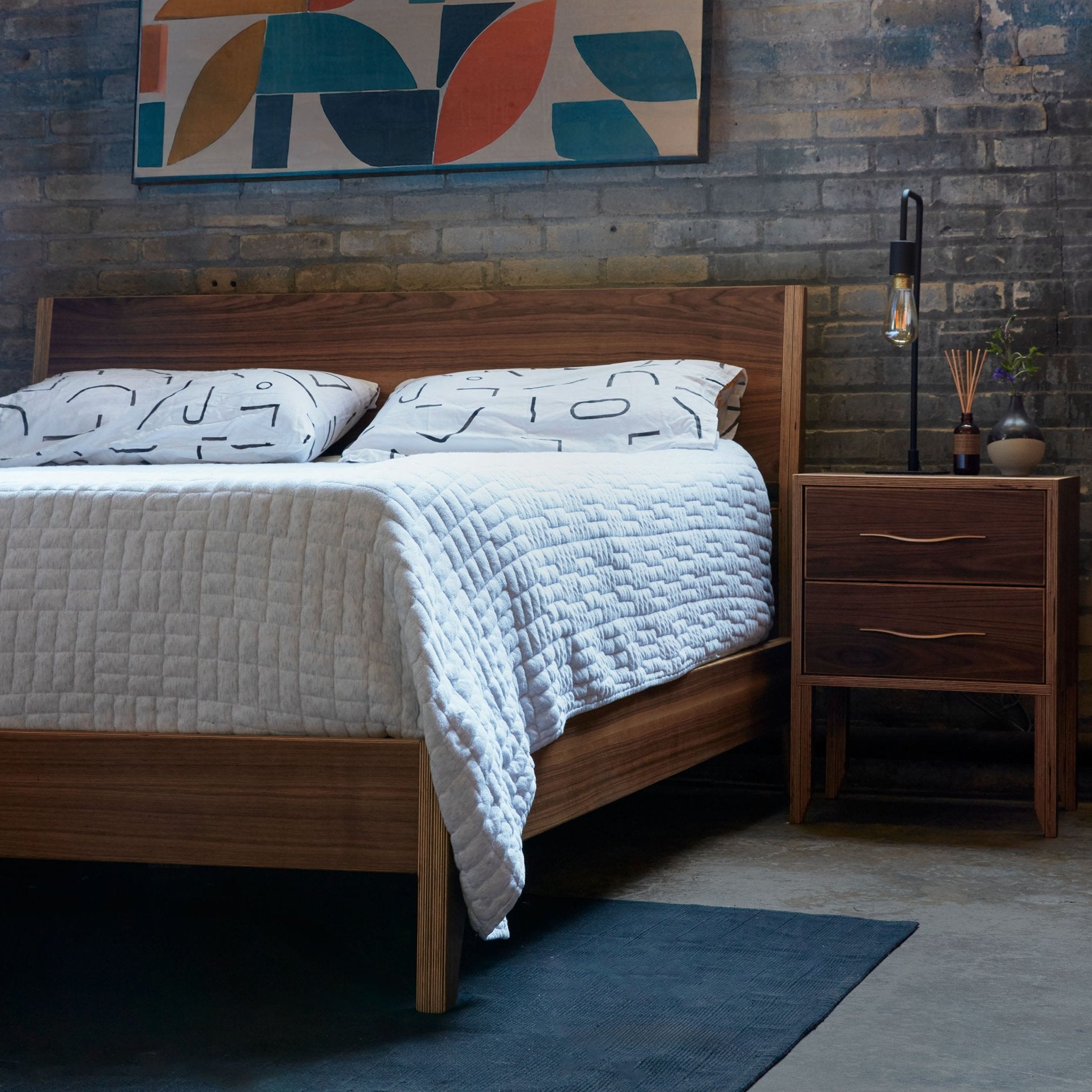 Walnut platform bed with matching nightstand, brick wall, patterned pillows and lamp