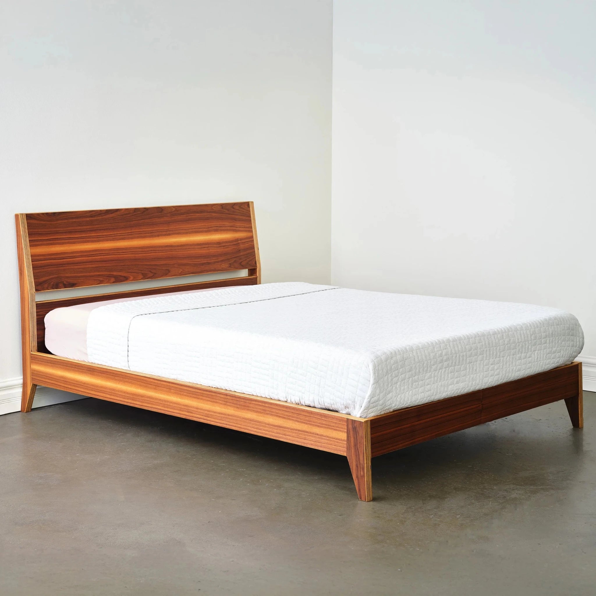 Walnut platform bed side view with angled headboard and white bedding, empty room