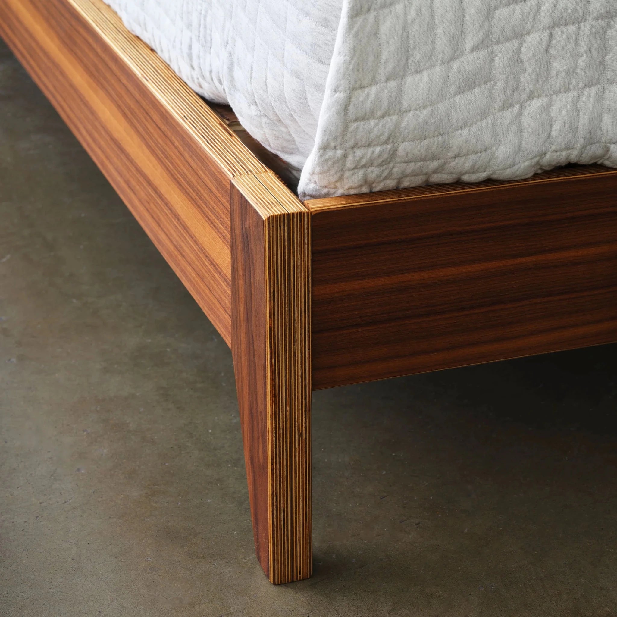 Close-up walnut platform bed foot leg showing layered birch edge, white quilt