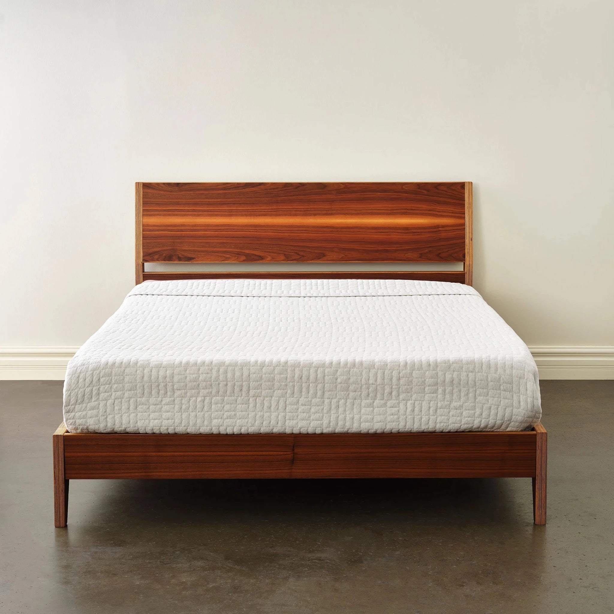 Walnut platform bed, front view, angled headboard, white quilted bedding, concrete floor