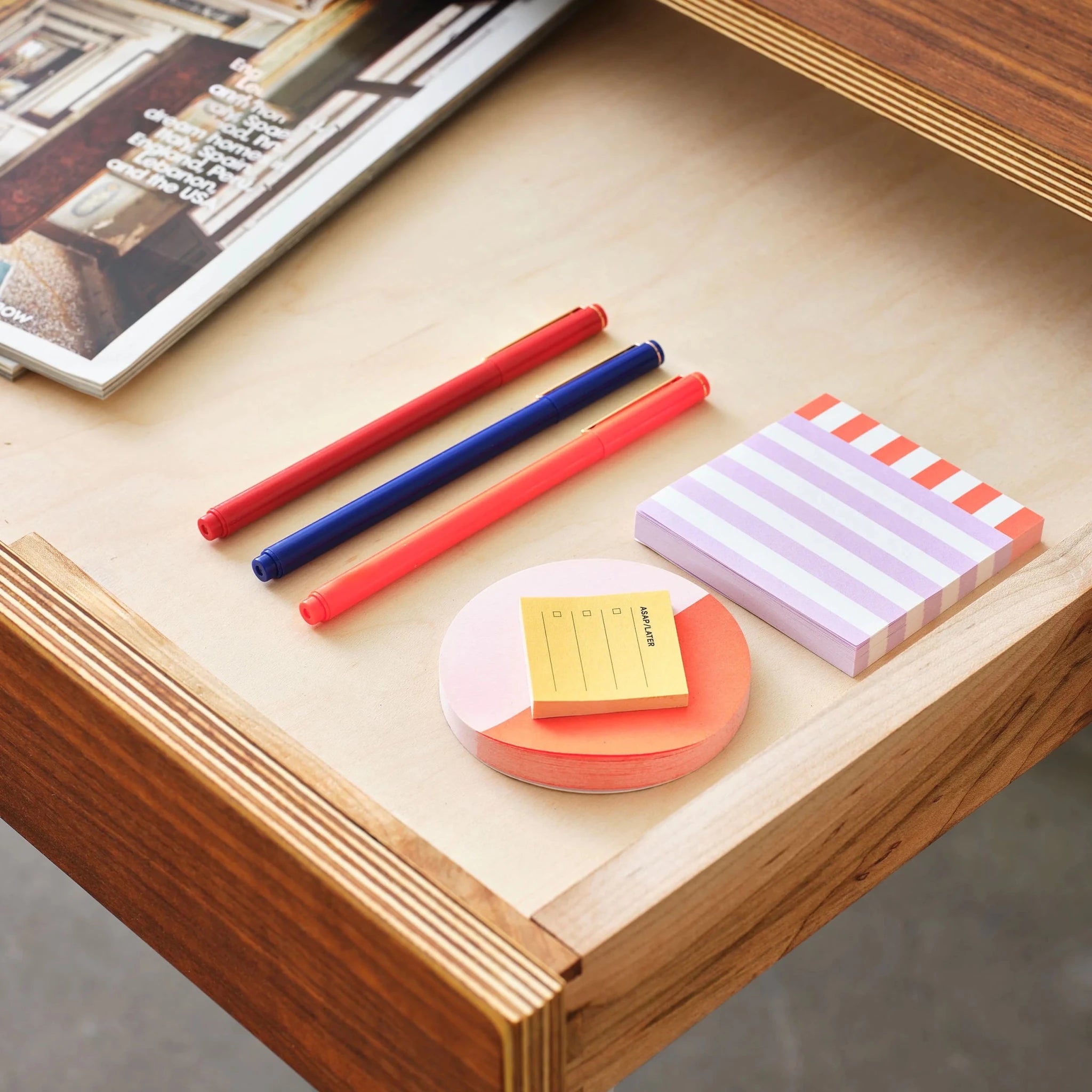 Open desk drawer with birch plywood interior holding colorful pens, sticky notes, magazine