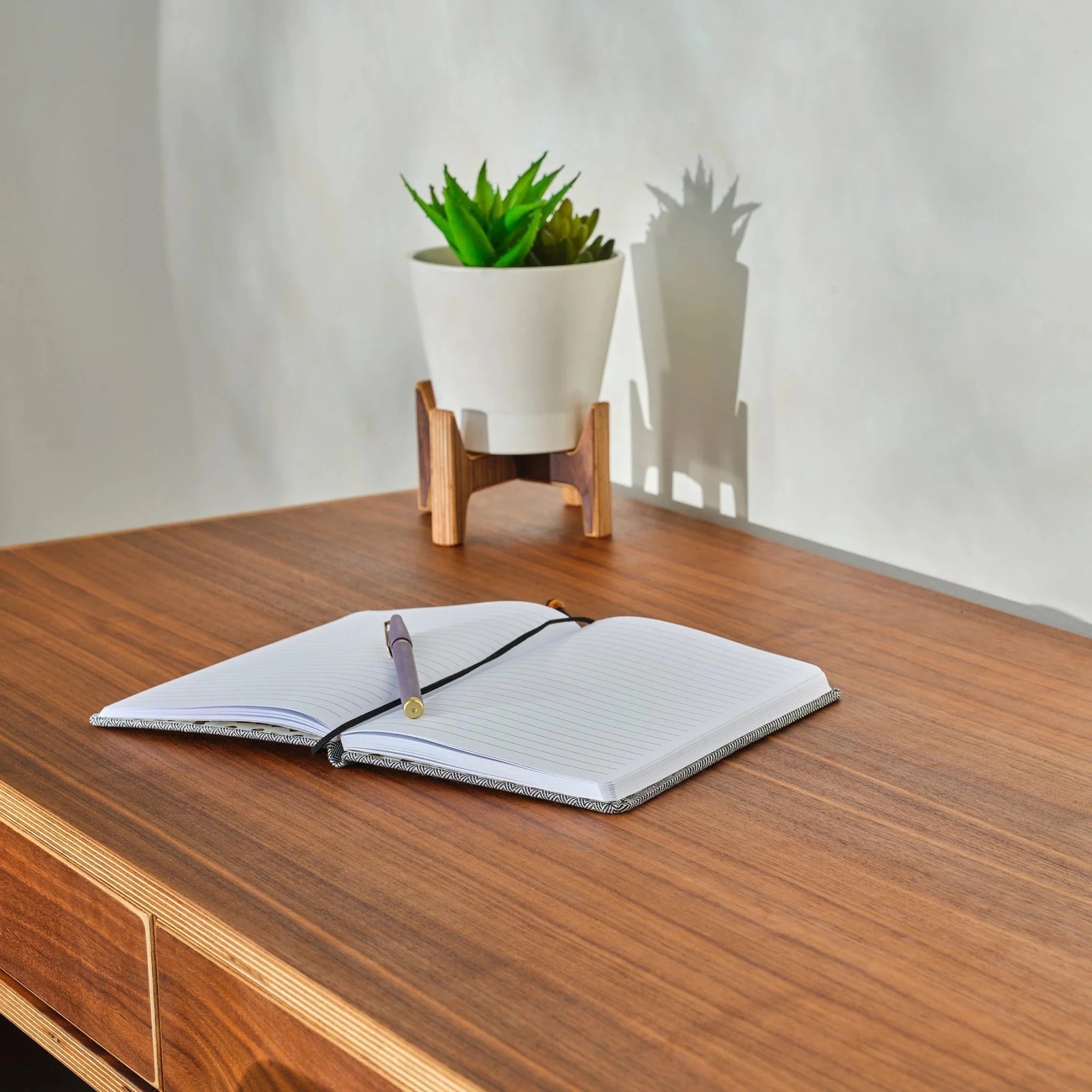 Small plant stand holding white potted succulent on walnut desk beside open notebook and pen