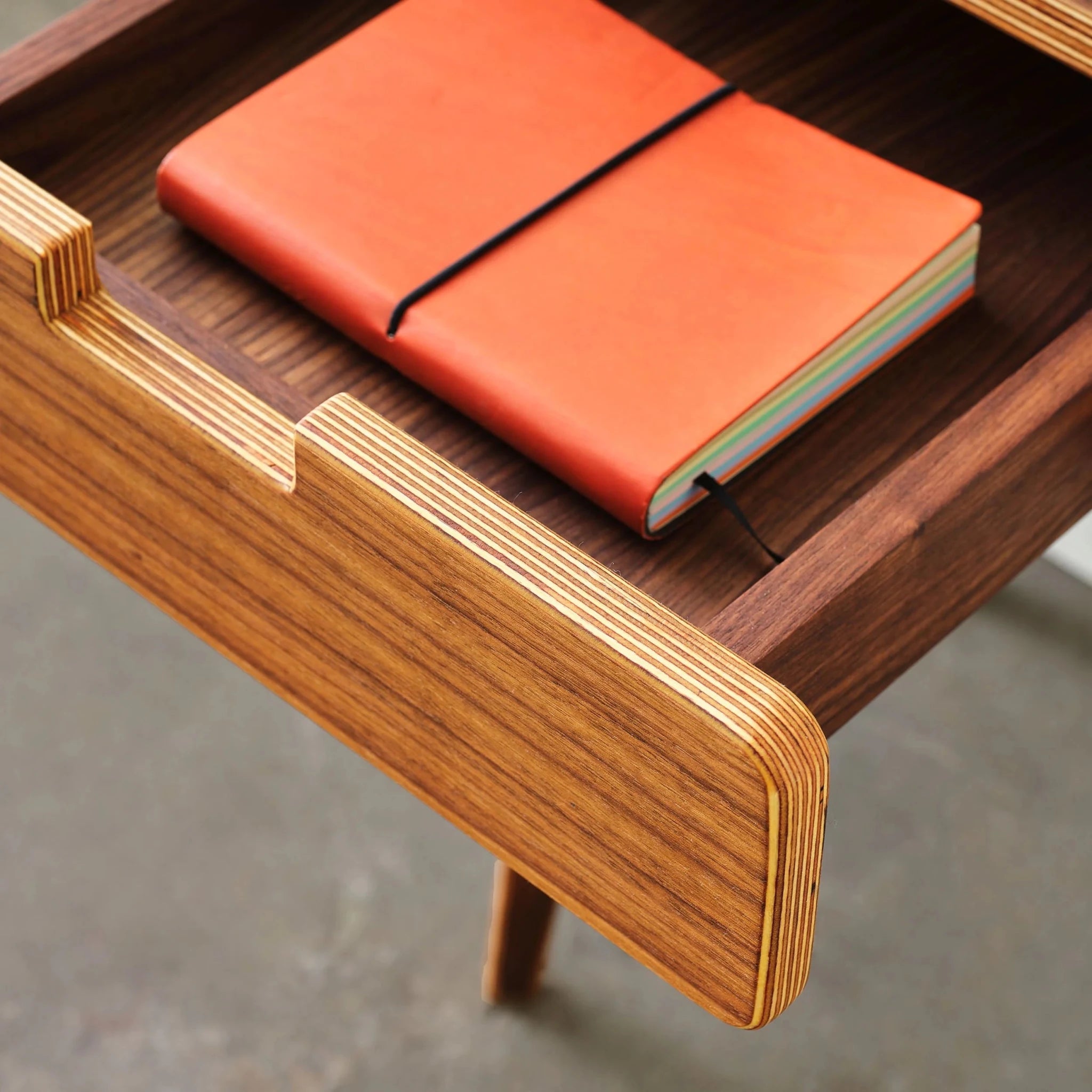 Open walnut end table drawer holding orange notebook, layered birch ply visible