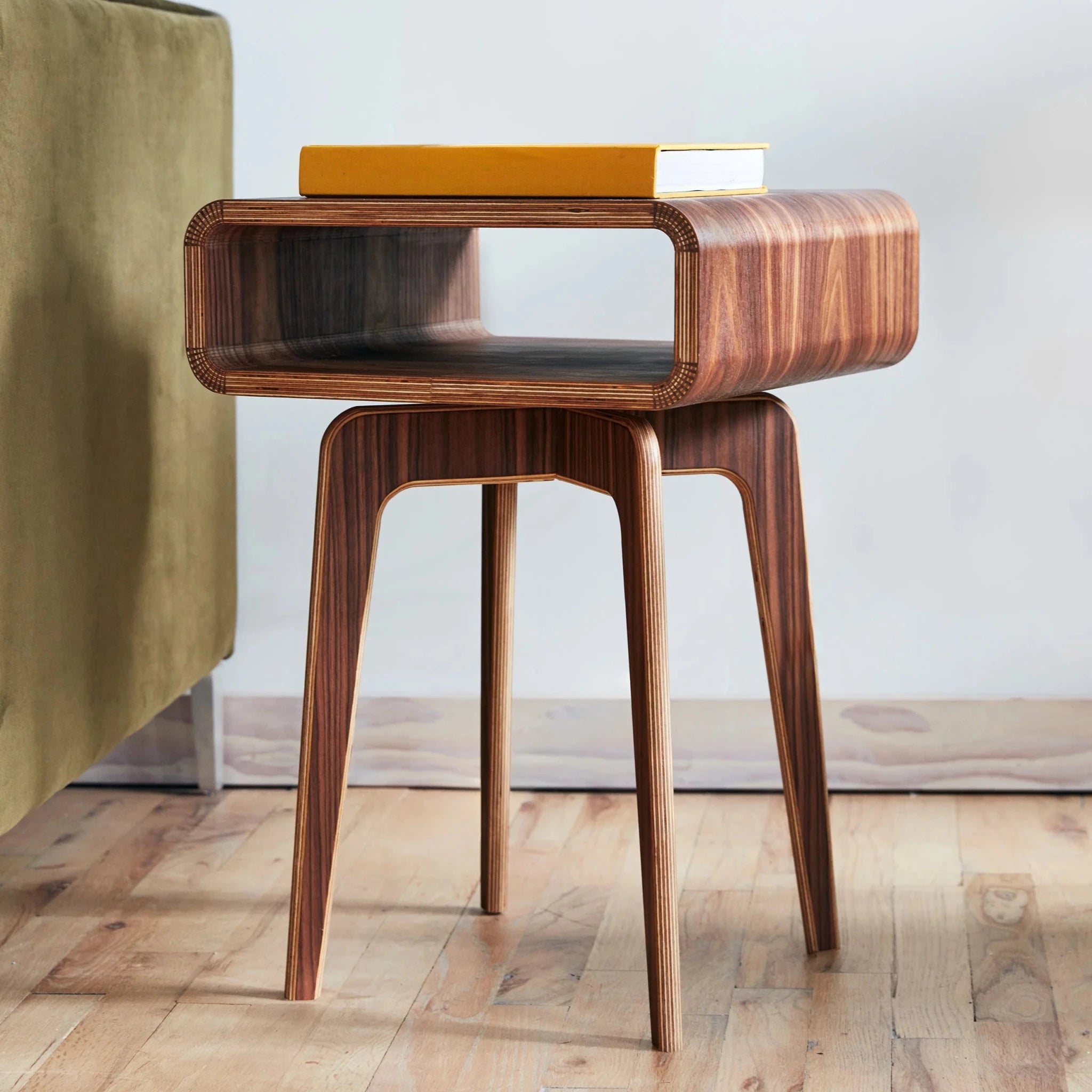 Walnut plywood end table beside olive sofa, open cubby, orange book on top