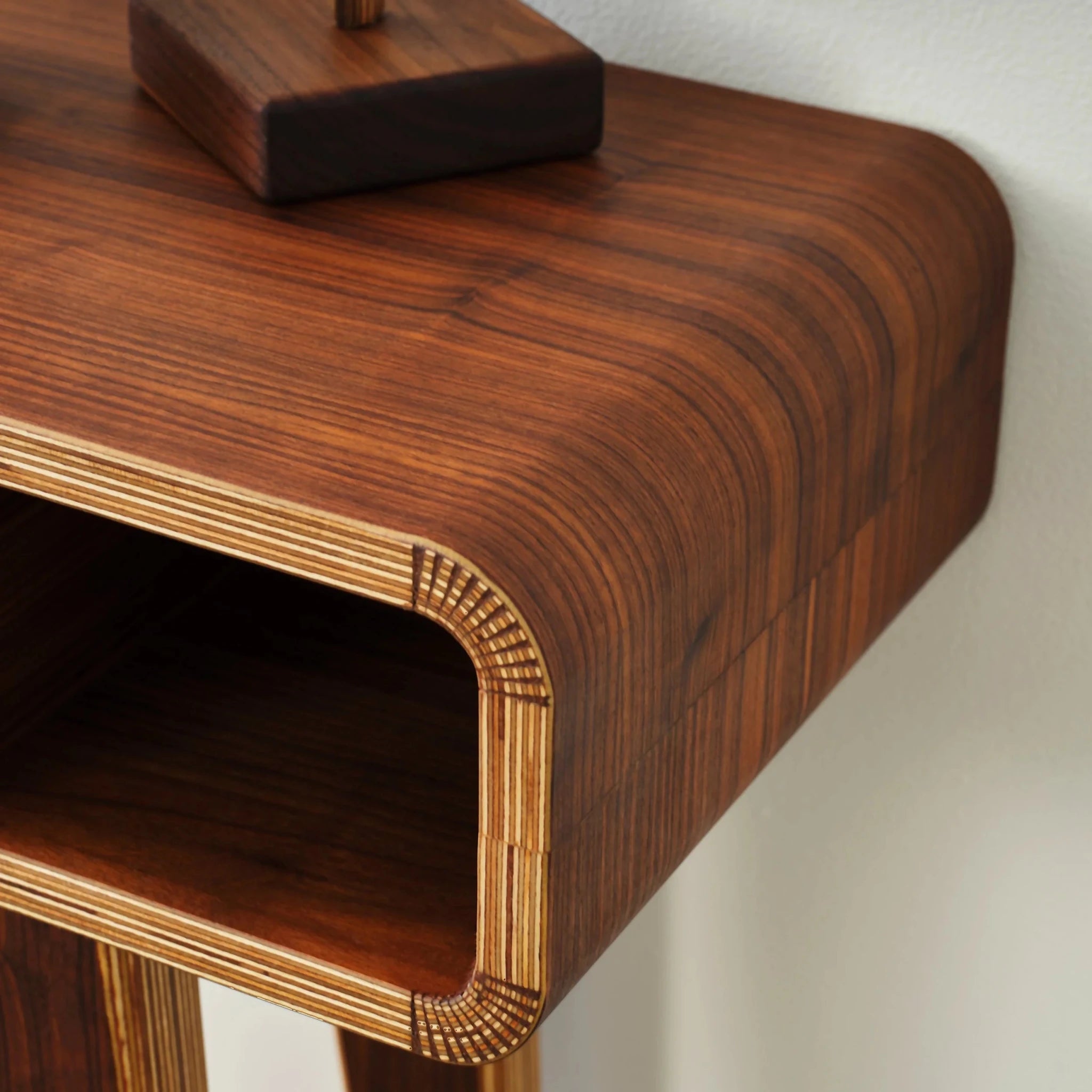 Overhead close-up of walnut tabletop corner, curved edge revealing layered birch plywood, sculpture base