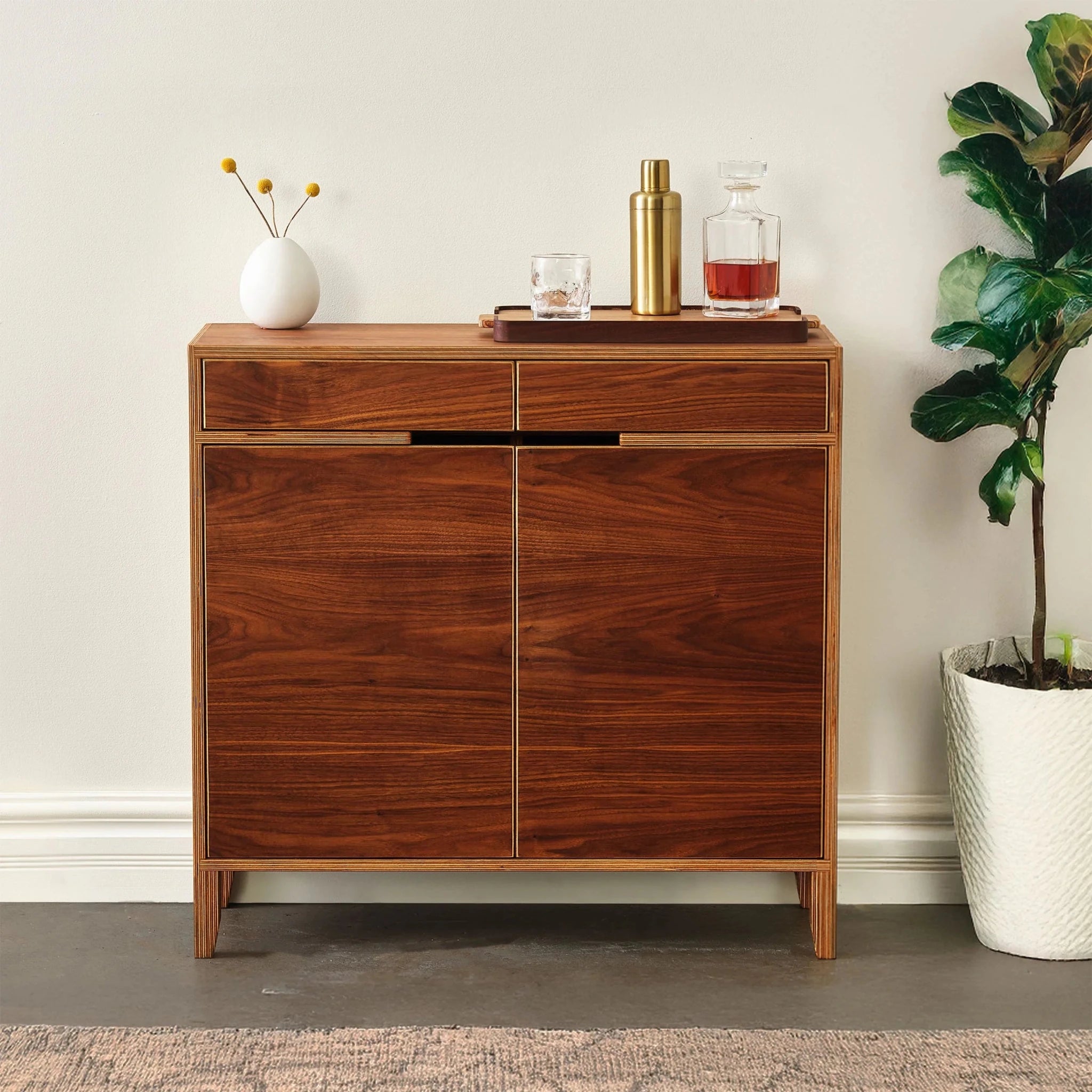 Walnut liquor cabinet, closed doors, styled with barware and fiddle-leaf fig plant