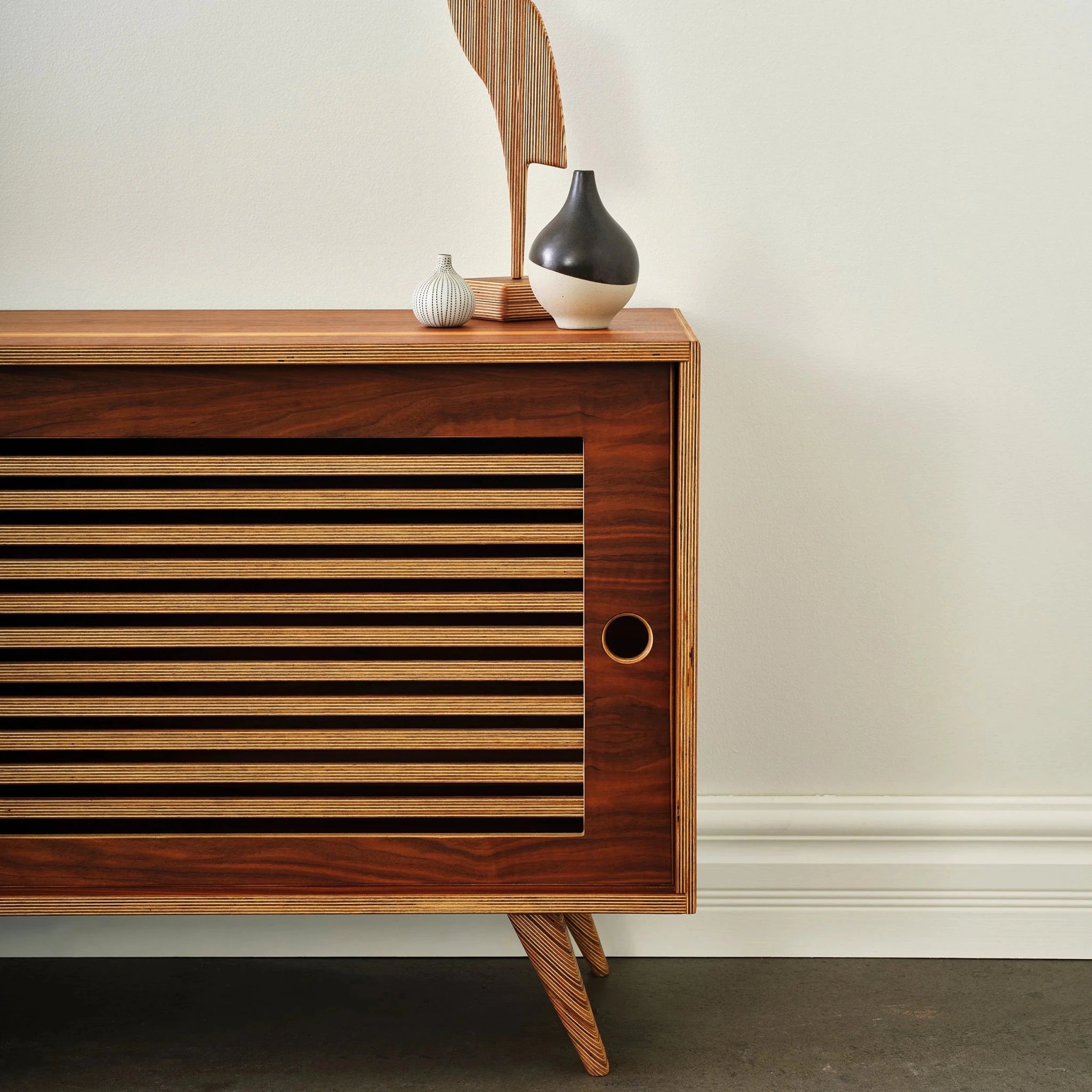 Close-up of walnut media console corner, slatted sliding door, birch ply edges, vases