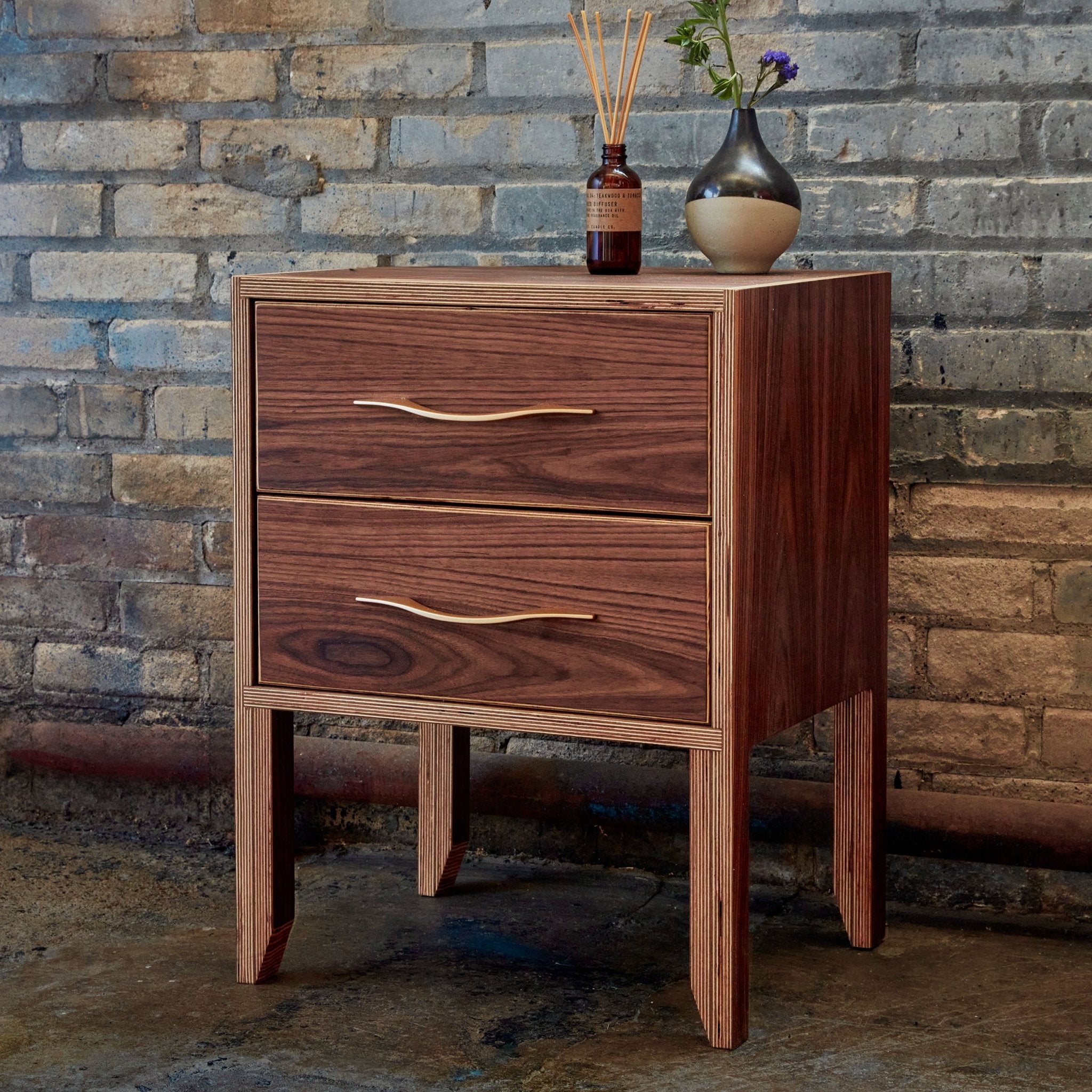 Walnut two-drawer nightstand against exposed brick, diffuser and bud vase with flower on top