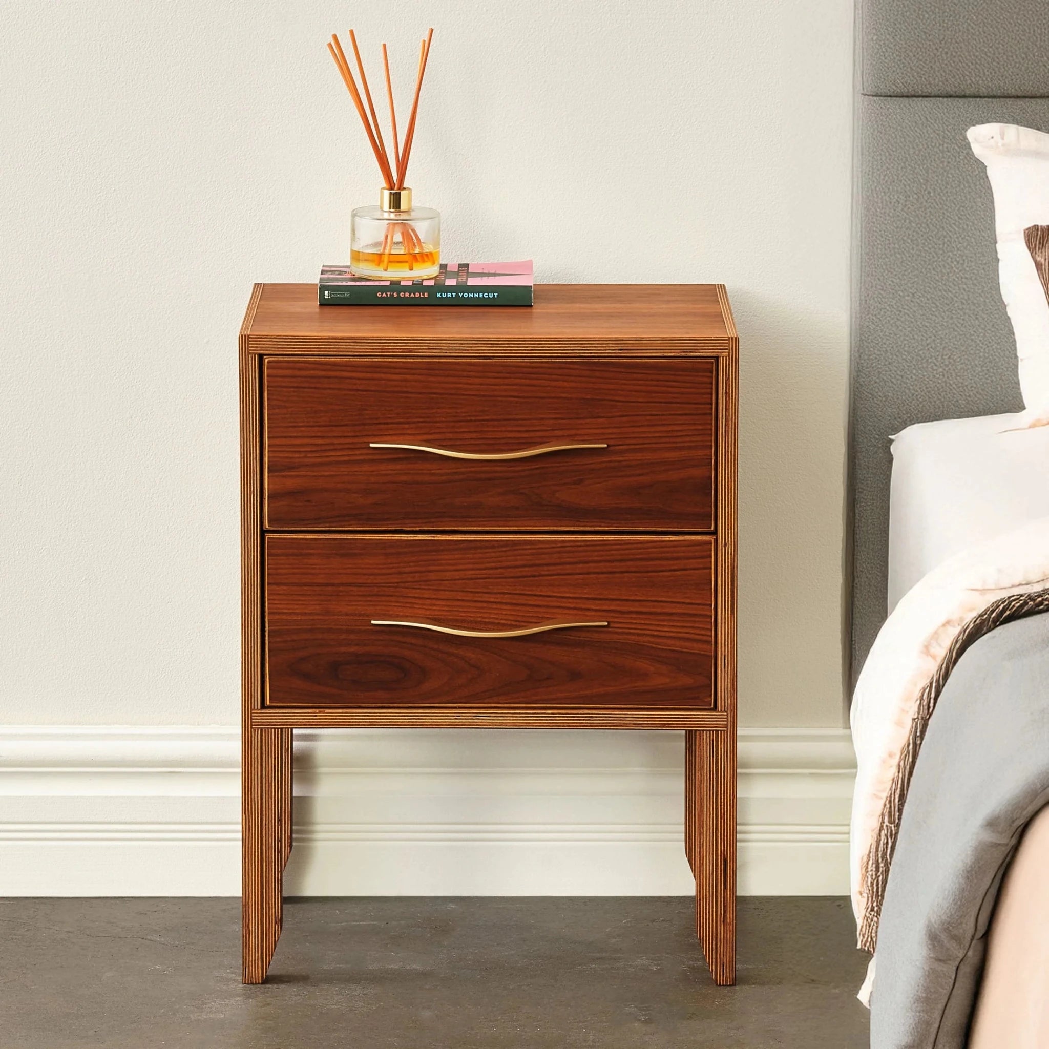 Walnut two-drawer nightstand with brass handles beside bed, reed diffuser and book on top