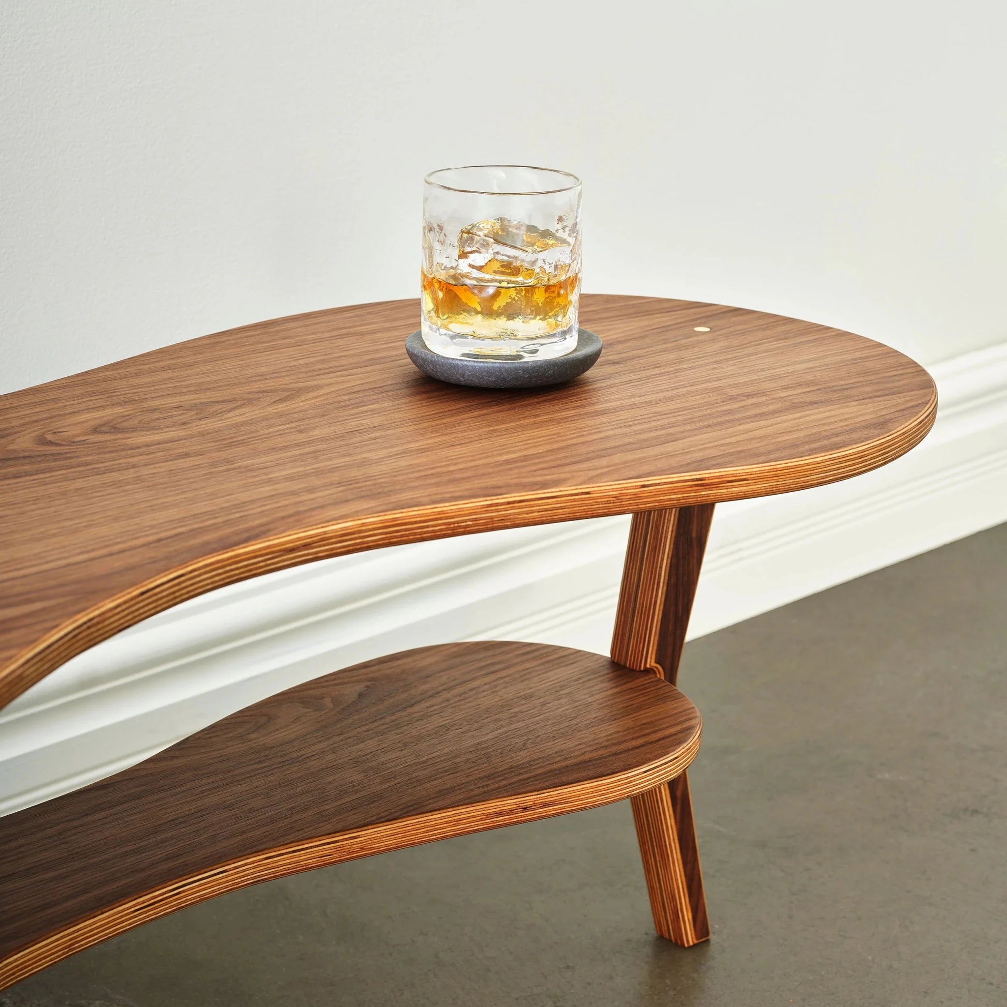 Partial view of walnut kidney cocktail table with whiskey glass, lower shelf, angled leg