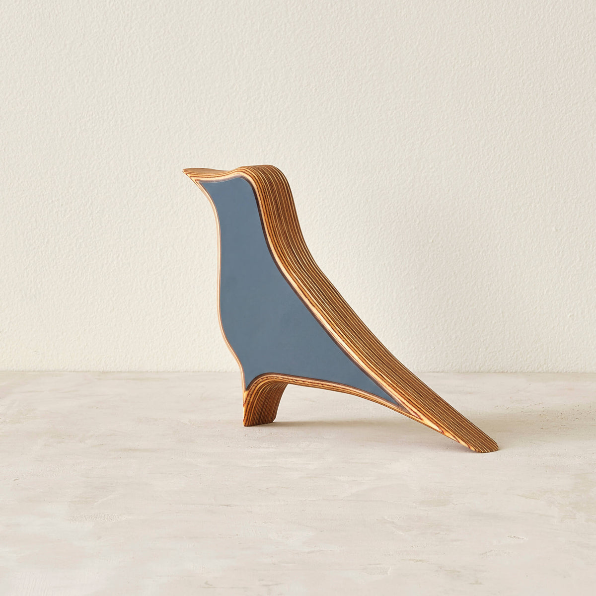 A modern bird-shaped decorative sculpture made of layered wood with a matte slate-blue laminate in a neutral space.