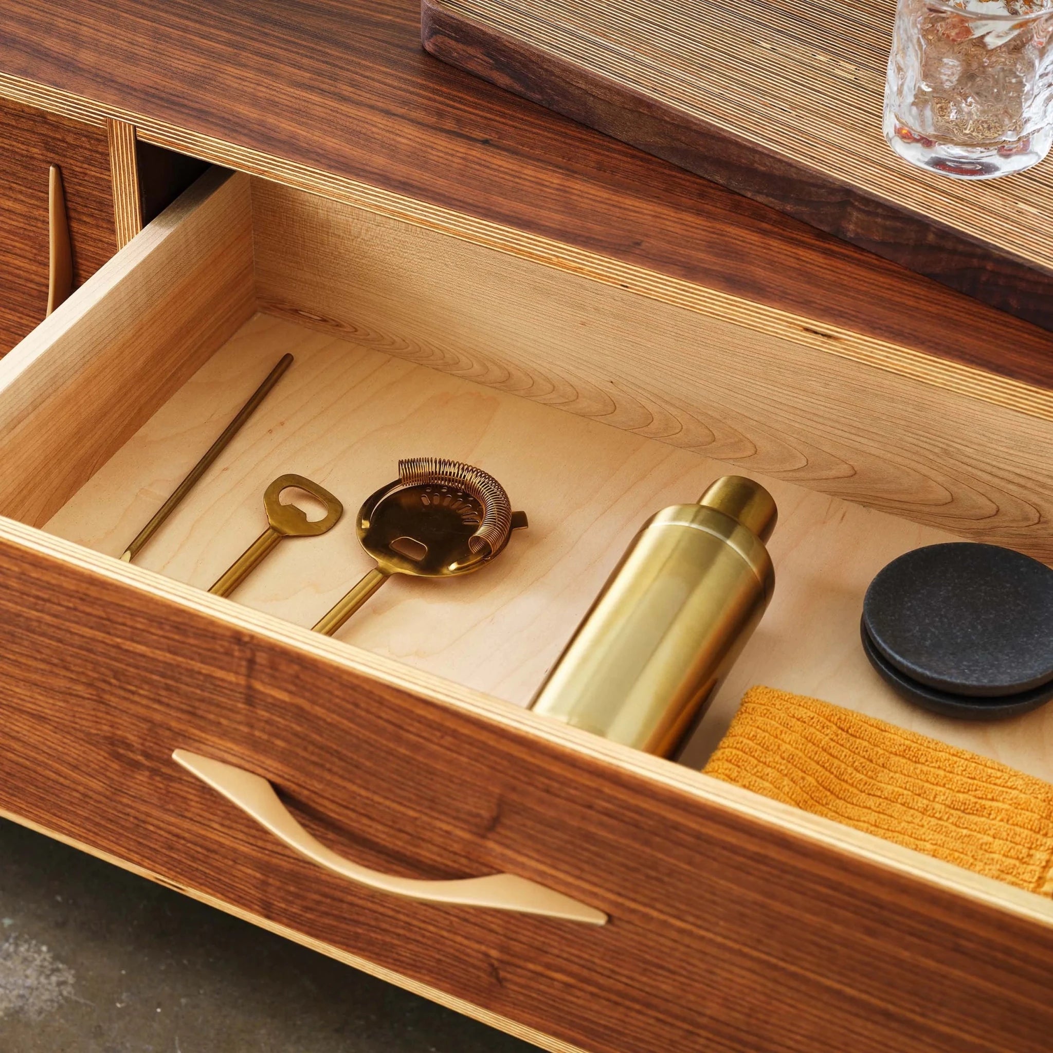 Open walnut sideboard drawer revealing brass barware, cocktail shaker and orange cloth inside