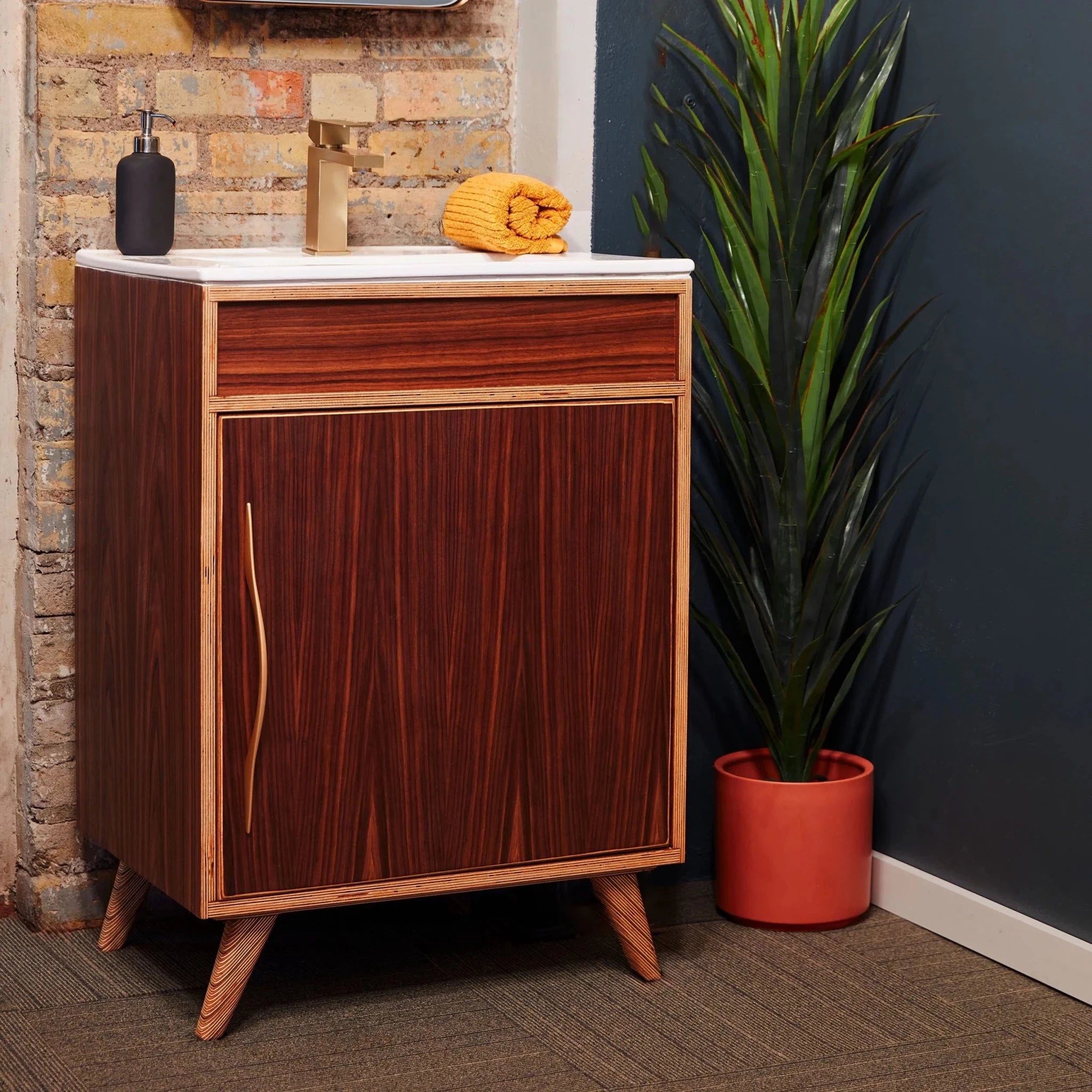 Angled rosewood single vanity, white top, brass faucet, potted plant, brick wall, splayed legs